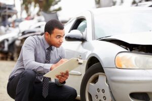 Driver photographs front-end damage on a car after a collision while holding an insurance policy folder.