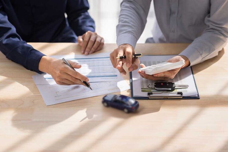 Driver reviewing an auto insurance policy beside a damaged car, repair estimate, and calculator on a desk.